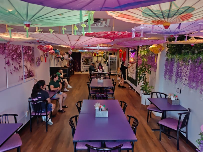 Restaurant interior with purple tables, colorful umbrellas on the ceiling, and customers dining.