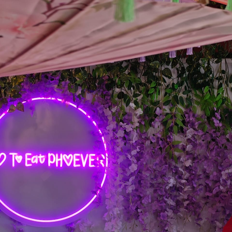 A purple neon sign saying "I Love To Eat Phoever" on a wall with green vines and purple flowers.