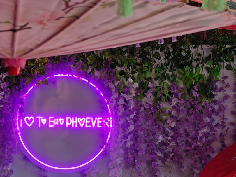 A purple neon sign saying "I Love To Eat Phoever" on a wall with green vines and purple flowers.